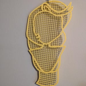 May include: A yellow, grid-like wall hanging in the shape of a person's head. The grid is made of small squares and the outline of the head is visible.