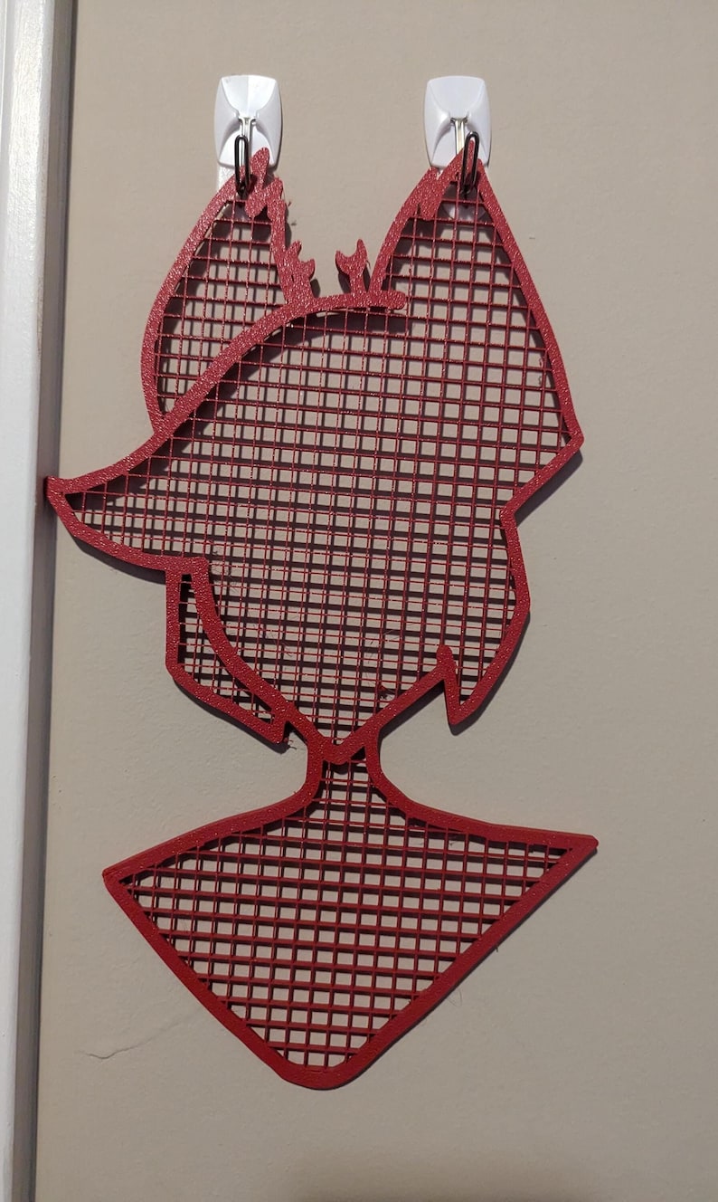 May include: Red metal wall decor in the shape of a person with a grid pattern. The decor is hanging on a wall with two white hooks.