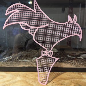 May include: A pink plastic rooster silhouette with a grid pattern. The rooster is standing on a small base.