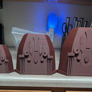 May include: Three brown decorative architectural models of varying sizes. Each model features a detailed facade with columns, arches, and stylized ornamentation. The models are arranged in a row on a countertop.