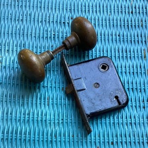 May include: Antique door hardware set featuring two oval-shaped bronze doorknobs connected by a rod, and a rectangular black metal mortise lock with a keyhole and bolt. The set is likely from a vintage door.