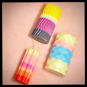 May include: Three colorful candles with different patterns and colors. The candles are arranged on a pink surface. The candles are made of wax and have a variety of colors, including yellow, purple, blue, pink, and orange.