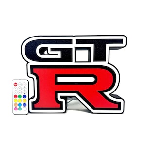 Op de afbeelding: Een zwart, rood en wit verlicht "GT-R" logo met een afstandsbediening. Het logo is een gestileerd ontwerp met de letters "GT" in zwart en "R" in rood. De afstandsbediening heeft meerdere kleuropties.