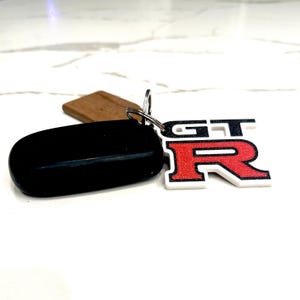 Pode incluir: Um chaveiro de carro preto e uma etiqueta de madeira presos a um chaveiro prateado. Um chaveiro com o logotipo GT-R branco e vermelho também está preso. O chaveiro tem formato oval.
