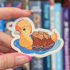 May include: A holographic sticker featuring a cartoon duck with a brown turtle shell body floating in blue water. The sticker has a shimmering star pattern overlay and a small pink heart icon near the duck's head, rendered in vibrant colors.