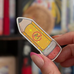 May include: A yellow pencil-shaped sticker with a holographic star pattern. The pencil features a red circle with a diagonal line through the letters AI in the center, a grey metal ferrule, and a pink eraser at the end with a white border.
