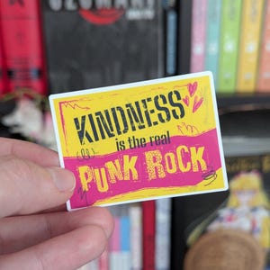 May include: A rectangular sticker with the text "KINDNESS is the real PUNK ROCK" in black and pink. The sticker has a yellow background with pink accents and small heart illustrations. The sticker is held in a person's hand.