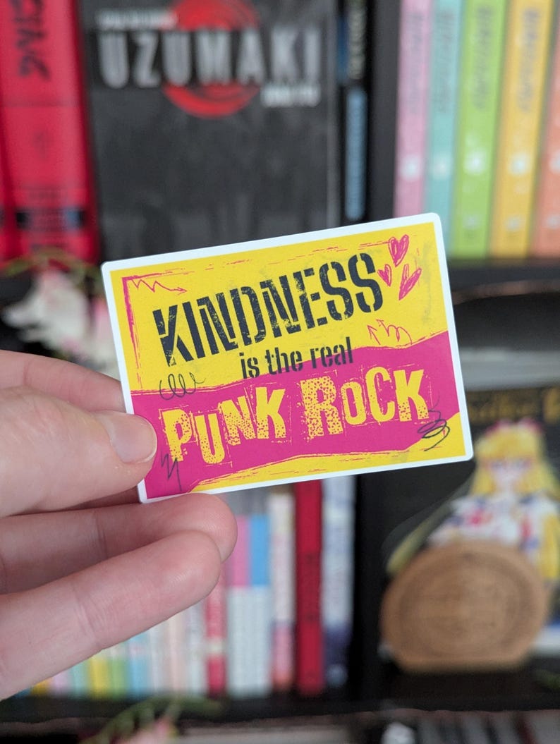 May include: A rectangular sticker with the text "KINDNESS is the real PUNK ROCK" in black and pink. The sticker has a yellow background with pink accents and small heart illustrations. The sticker is held in a person's hand.