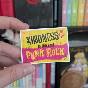 May include: A rectangular sticker with a yellow and pink background. The sticker has the words "KINDNESS is the real PUNK ROCK" in black and white text. Two small pink hearts are in the upper right corner.