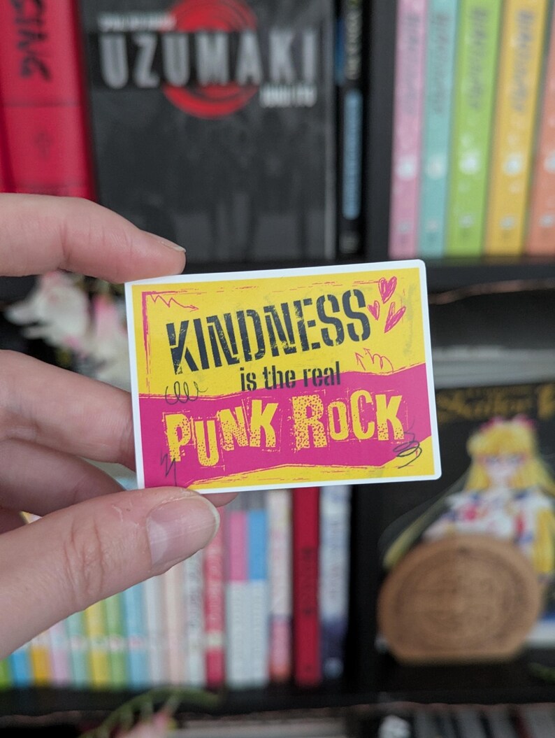 May include: A rectangular sticker with a yellow and pink background. The sticker has the words "KINDNESS is the real PUNK ROCK" in black and white text. Two small pink hearts are in the upper right corner.