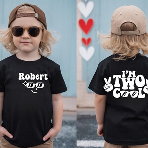 May include: A black t-shirt with the name "Robert" and sunglasses graphic on the front. The back of the shirt says "I'm Two Cool" with peace signs. The child is wearing a brown cap and sunglasses.