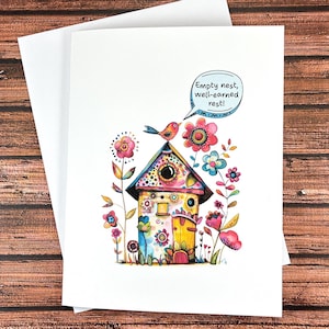 May include: A white greeting card featuring a colorful watercolor illustration of a birdhouse surrounded by flowers. A small bird sits on the roof with a speech bubble containing the text: Empty nest, well-earned rest! A plain white envelope is behind.