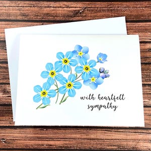 May include: A white sympathy card with a watercolor illustration of blue forget-me-not flowers with yellow centers. The card includes the text "with heartfelt sympathy" in a black script font. The card is paired with a white envelope.