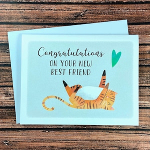 May include: Greeting card with a light blue background and the words "Congratulations ON YOUR NEW BEST FRIEND". The card features an illustration of a tiger with orange and black stripes. A green heart is above the tiger.