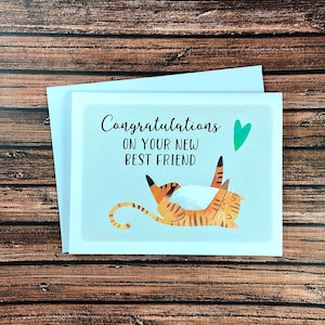 May include: A greeting card with the text "Congratulations on your new best friend" and a cartoon illustration of a playful orange and brown striped cat. A green heart is above the cat. The card is on a white envelope.