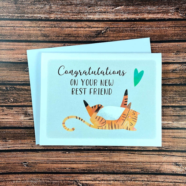 May include: A greeting card with the text "Congratulations on your new best friend" and a cartoon illustration of a playful orange and brown striped cat. A green heart is above the cat. The card is on a white envelope.