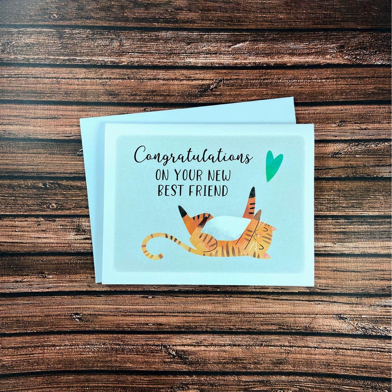May include: Greeting card with a light blue background and the words "Congratulations ON YOUR NEW BEST FRIEND". The card features an illustration of a tiger with orange and black stripes. A green heart is above the tiger.
