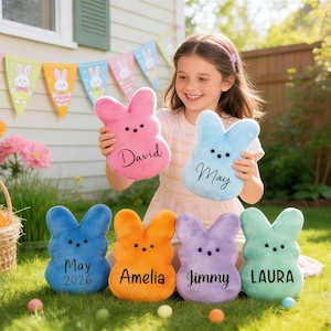 Puede incluir: Una niña sostiene dos almohadas de felpa con forma de conejito, una rosa y otra azul, cada una con un nombre bordado. Otras almohadas de conejito en azul, naranja, morado y verde están en la hierba, cada una con un nombre. Decoraciones temáticas de Pascua en el fondo.