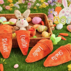 May include: An Easter-themed display featuring plush carrot-shaped pouches with names embroidered on them, a stuffed bunny, and decorated Easter eggs. The pouches are orange with green tops, and the names are in white. A wooden Easter bunny cutout is in the background.