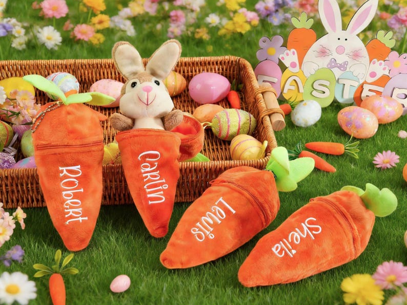 May include: An Easter-themed display featuring plush carrot-shaped pouches with names embroidered on them, a stuffed bunny, and decorated Easter eggs. The pouches are orange with green tops, and the names are in white. A wooden Easter bunny cutout is in the background.
