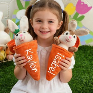May include: A smiling girl holds two plush bunny toys in orange carrot-shaped holders. The bunnies have brown and white fur, with pink noses and long ears. The carrot holders have the names "Kaylor" and "Isabel" written on them.
