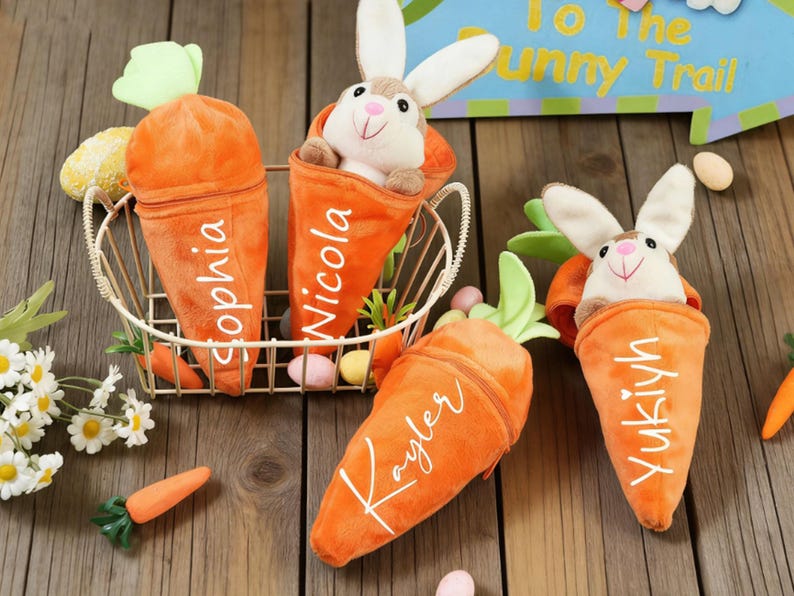 May include: Orange plush carrot-shaped containers with white lettering, each holding a stuffed bunny. The carrots are in a gold wire basket, with decorative carrots and flowers. Easter-themed gift.