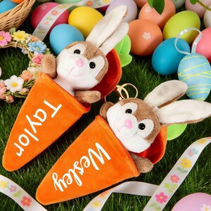May include: Two plush bunny keychains emerge from orange carrot-shaped holders, one labeled "Taylor" and the other "Wesley." The scene is set on green grass, surrounded by colorful Easter eggs, ribbons, and a floral wreath, creating a festive holiday theme.
