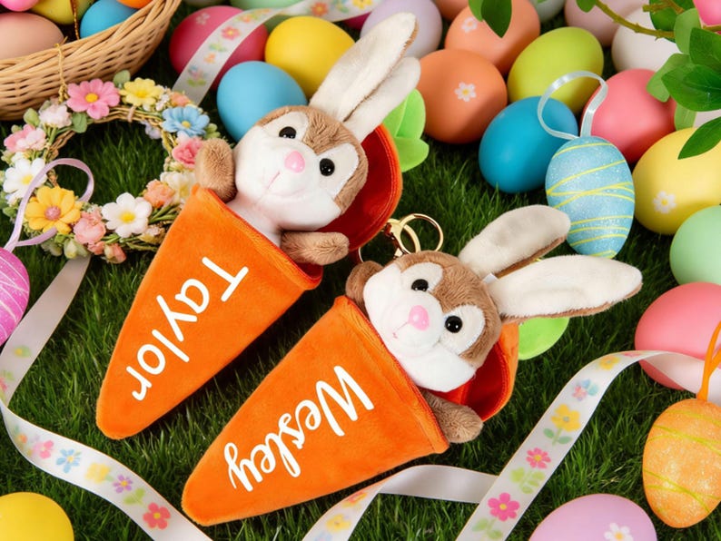 May include: Two plush bunny keychains emerge from orange carrot-shaped holders, one labeled "Taylor" and the other "Wesley." The scene is set on green grass, surrounded by colorful Easter eggs, ribbons, and a floral wreath, creating a festive holiday theme.