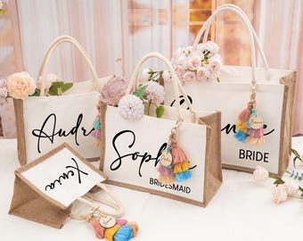 May include: Four white canvas tote bags with brown jute trim and personalized names. The bags have colorful tassel keychains with wooden tags. The names on the bags are "Bride", "Audra", "Sophia", and "Kiana".