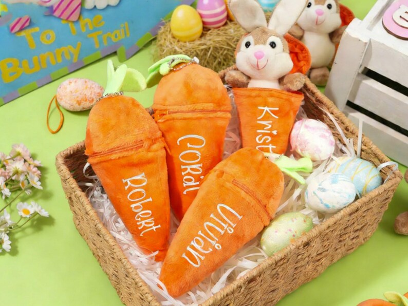 May include: A woven basket filled with plush carrot-shaped pouches, each with a name embroidered on it. The basket also contains decorative Easter eggs and a stuffed bunny. The pouches are orange with green tops.