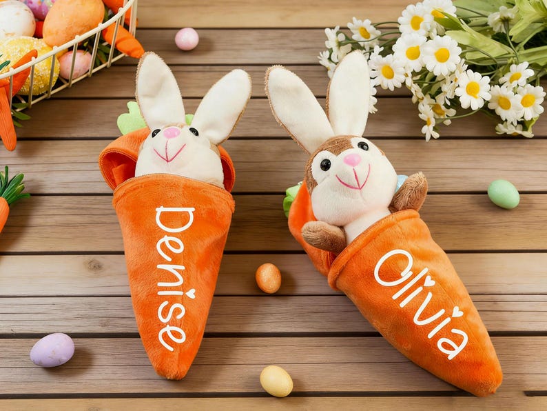May include: Two plush Easter bunny toys in orange carrot-shaped holders. One bunny is white and the other is brown. The carrots have names printed on them. Easter eggs and flowers are in the background.