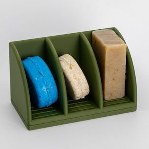 May include: A green soap dish with three compartments. The compartments hold a blue soap bar, a white soap bar, and a rectangular bar of soap.