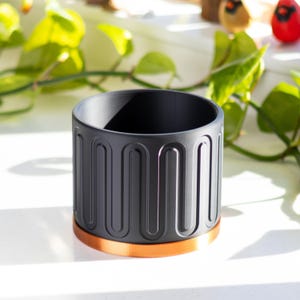 May include: A black cylindrical planter with a copper-colored rim. The planter has a textured, geometric design.