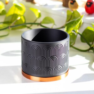 May include: Black planter with a geometric pattern and a copper-colored rim. The planter is made of ceramic and has a modern design.