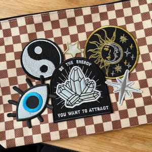 May include: A collection of embroidered patches with celestial and spiritual themes. Includes a yin-yang symbol, an eye, a sun and moon design, a crystal, and star shapes. The patches are on a brown and tan checkered background.