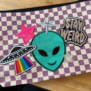 May include: A zippered pouch with a purple and white checkered pattern. The pouch is decorated with embroidered patches, including a green alien head, a UFO with a rainbow beam, and the words "Stay Weird".