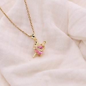 Puede incluir: Collar dorado con un colgante con una gema rosa en forma de corazón atravesada por una espada. El corazón está rodeado por un halo de pequeñas piedras transparentes. La cadena y el colgante son de metal brillante color oro.
