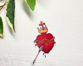 May include: A sticker featuring a red heart pierced by an arrow, with a crown of thorns and a cross above. The heart has flames around it and is dripping with blood. The sticker is on a white background.