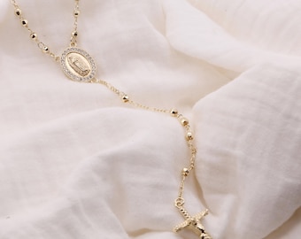 Puede incluir: Collar de rosario dorado con un colgante central ovalado que representa una figura religiosa, rodeado de pequeñas piedras transparentes. El collar tiene una cadena de cuentas y un colgante de crucifijo.