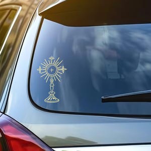 Puede incluir: Una calcomanía de vinilo dorada de una custodia, un símbolo religioso, está adherida a la ventana de un automóvil. La custodia presenta una cruz central dentro de un diseño de rayos de sol, con detalles intrincados y una base de pedestal.