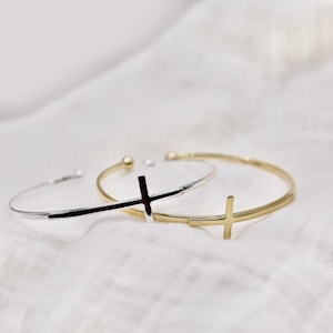 May include: Two open cuff bracelets with cross designs. One bracelet is silver-toned, and the other is gold-toned. The bracelets are displayed on a white, textured surface. The cross is positioned on the side of the bracelets.