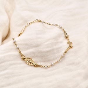 Pode incluir: Pulseira de rosário dourada com um berloque de cruz e um pingente oval com uma figura religiosa. A pulseira é adornada com pequenas pérolas brancas. O design é delicado e elegante, adequado para ocasiões religiosas ou uso diário.