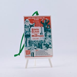 May include: A miniature Christmas ornament featuring a movie poster for "Santa Claus Conquers the Martians." The poster is red, white, and blue, mounted on a tiny white easel with a green string for hanging. The text on the poster reads "Santa Brings Christmas Fun to Mars!"