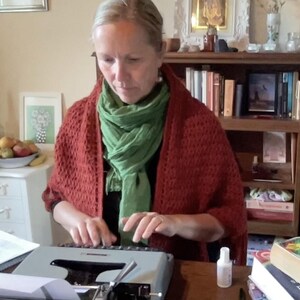 Puede incluir: Una persona viste un chal de ganchillo color óxido sobre una prenda negra y una bufanda verde brillante, escribiendo en una máquina de escribir manual gris claro. La máquina tiene un teclado negro y una palanca de carro metálica sobre un escritorio.