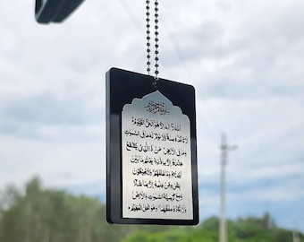 May include: A black rectangular car charm with a silver-colored religious text. The charm is suspended by a beaded chain. The text is in Arabic script and is centered on the charm.