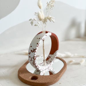 Pode incluir: Um vaso de cerâmica branco com manchas marrons, em forma de anel, fica sobre uma base de cortiça com um espelho. Há flores brancas secas no vaso.