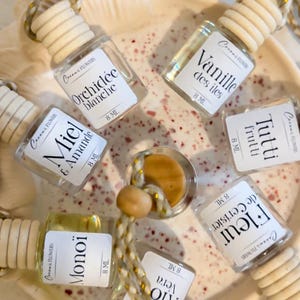 Può includere: Una collezione di piccole bottiglie di vetro con tappi di legno, ciascuna etichettata con un nome di fragranza diverso. Le bottiglie sono disposte su una superficie decorativa maculata e sono collegate da un cordoncino decorativo. Le etichette includono "Orchidée Blanche" e "Vanille des Iles".