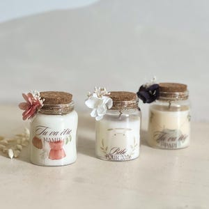 May include: Three small glass jars with cork lids, each decorated with a small flower and a label. The jars contain white wax candles. The labels feature French text and illustrations. One jar has a pink flower, another a white flower, and the last a dark purple flower.