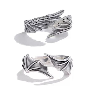 May include: Two silver rings. The top ring has a wing design with detailed feather textures. The bottom ring features a bat wing design with pointed tips. Both rings are open-ended, allowing for adjustable sizing.