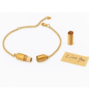 May include: Gold-colored message bracelet with a cylindrical pendant that opens to reveal a small scroll. A separate scroll and a small card with "I Love You" written on it are also present.